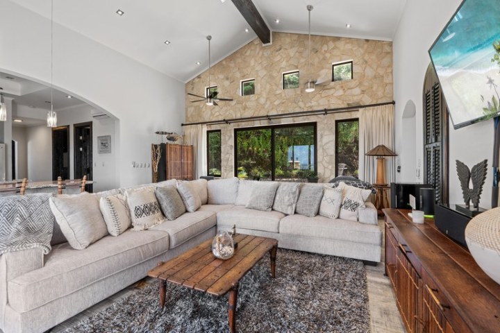 Spacious living room with beige sectional, wood table, large TV, and stone wall with windows.