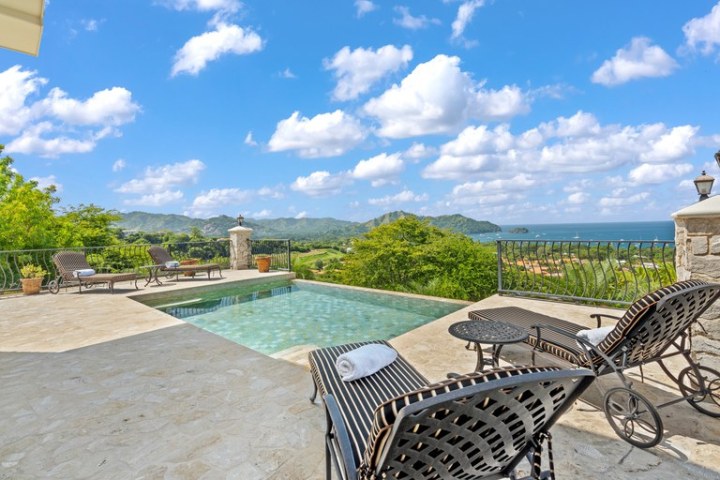 Patio with lounge chairs, pool, and scenic view of mountains and ocean under a cloudy sky.