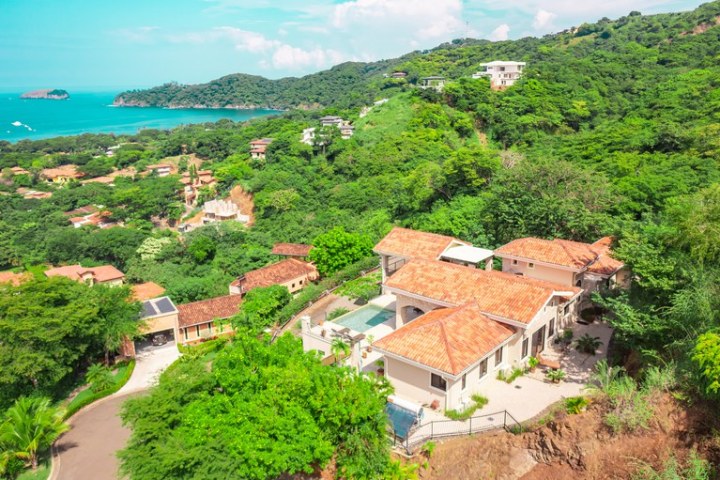 Aerial view of a house with a pool, surrounded by lush greenery and a distant view of the ocean.
