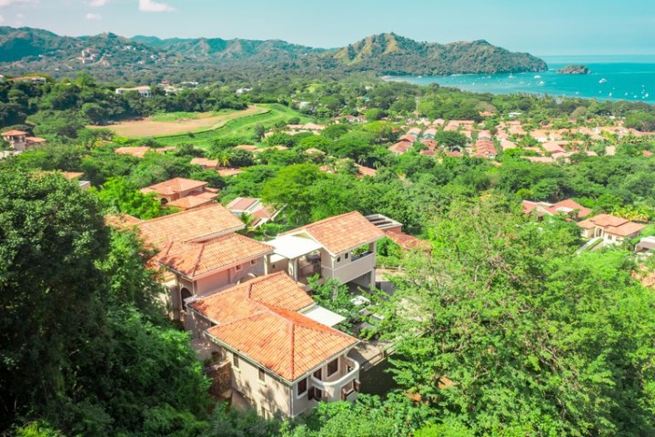 Aerial view of houses with red roofs, lush greenery, and distant ocean.