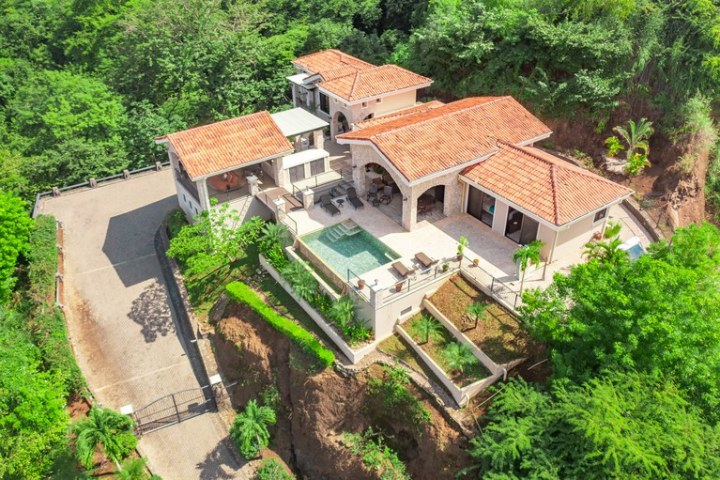 Aerial view of a hillside villa with a pool, surrounded by lush greenery.