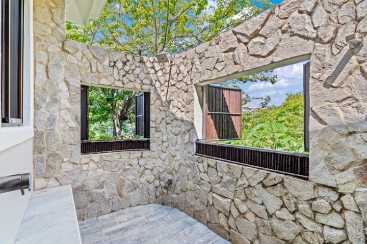 Stone-walled outdoor shower with two openings, surrounded by trees.