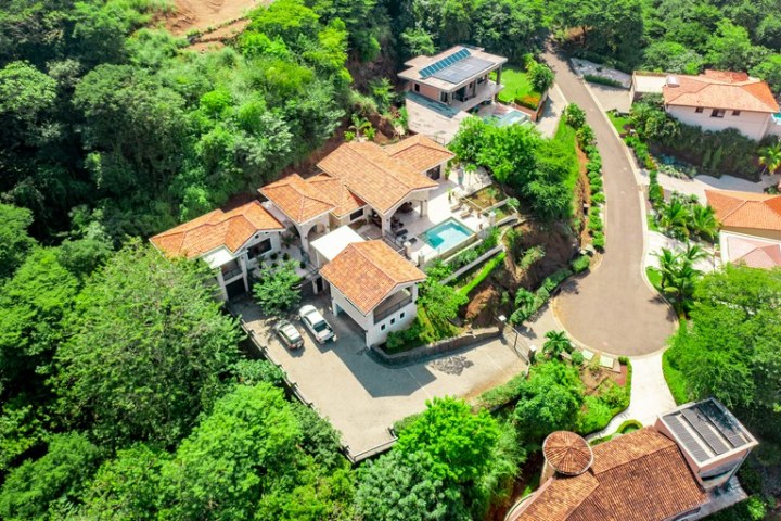 Aerial view of a large villa with a pool, surrounded by trees and a winding road.