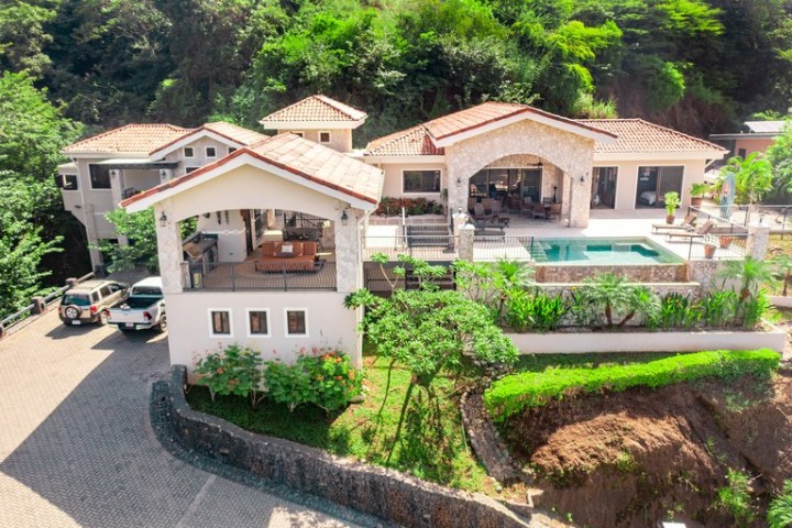 Large house with pool, surrounded by trees and parked cars, aerial view.