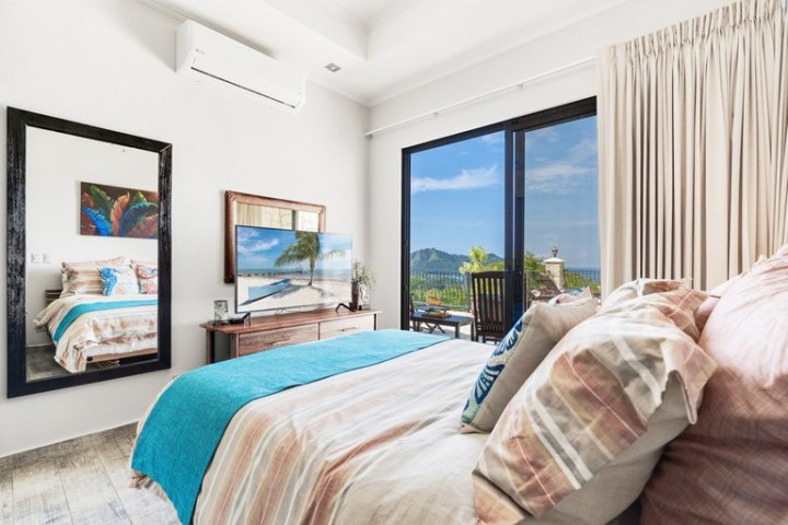 Bright bedroom with a bed, TV, mirror, and balcony with scenic view.