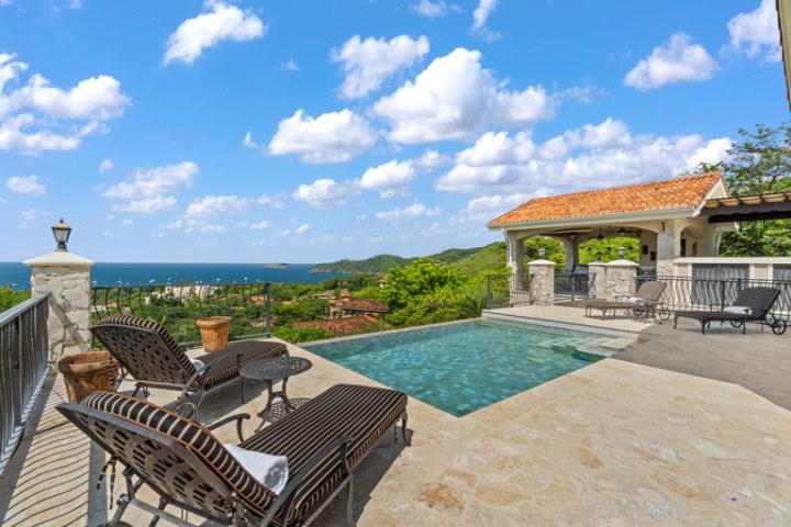 Infinity pool with lounge chairs, ocean view, and pavilion under clear blue sky.