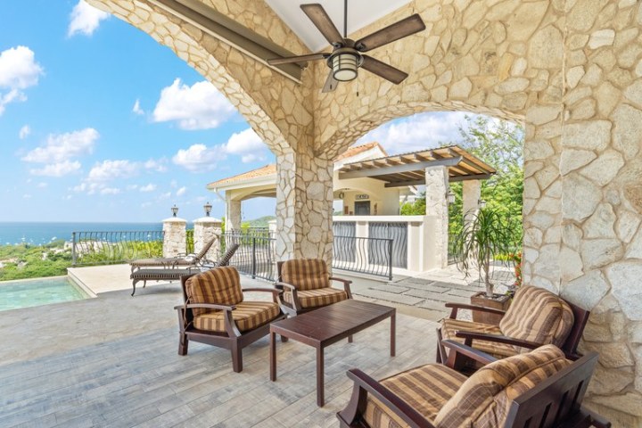 Outdoor patio with stone walls, ceiling fan, chairs, table, and ocean view.