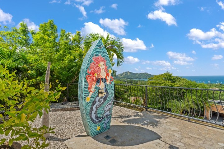Mosaic surfboard with mermaid art near railing and green trees, ocean view.