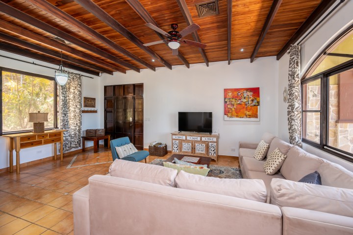 Cozy living room with beige sofa, TV, ceiling fan, wooden ceiling beams, and large windows.