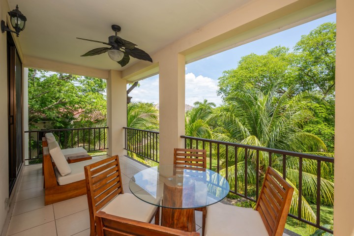 Outdoor balcony with wooden chairs, glass table, and view of lush green trees.