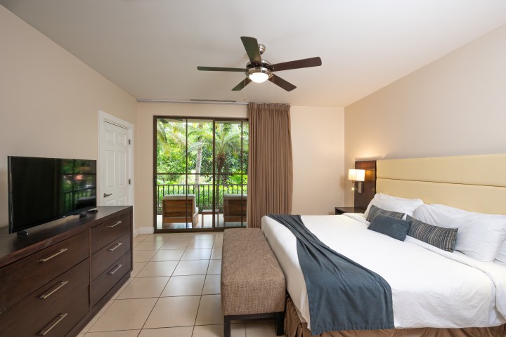 Modern bedroom with king bed, TV, dresser, and balcony view of tropical greenery.