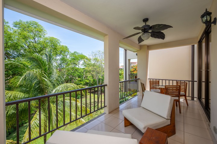 Covered balcony with chairs, table, and ceiling fan, overlooking lush greenery.