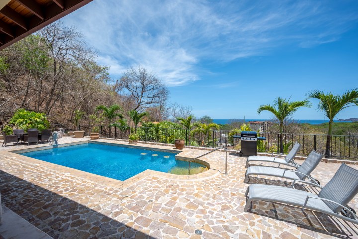 Outdoor pool with lounge chairs and a grill, surrounded by trees and overlooking a distant ocean view.