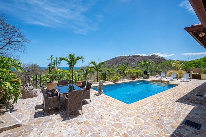 Outdoor pool area with lounge chairs and dining table overlooking hilly landscape.