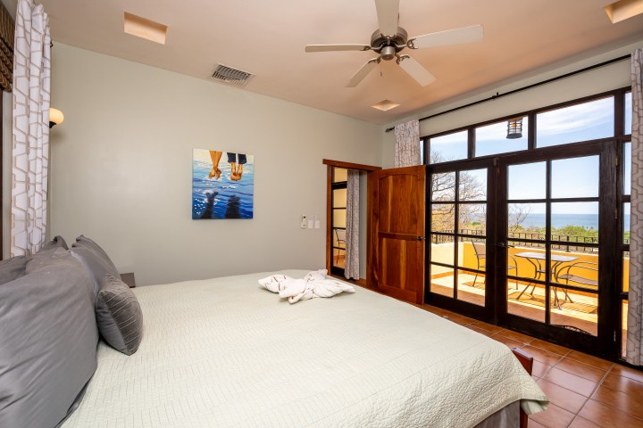 Bedroom with bed, ceiling fan, painting, and balcony overlooking a scenic view.