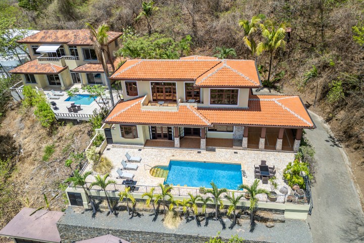 Aerial view of two villas on a hillside, each with red roofs and pools surrounded by tropical greenery.