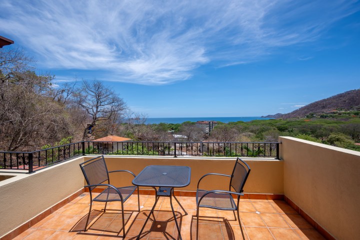 Terrace with two chairs and table overlooking ocean and hills under a blue sky.