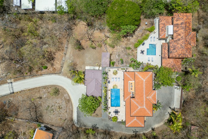 Aerial view of a house with an orange roof, two pools, trees, and a road.