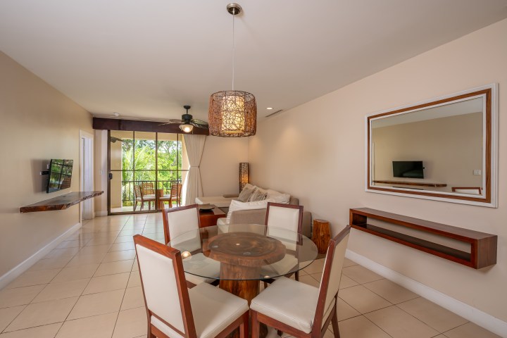 Modern living and dining area with glass table and beige sofa, leading to a balcony with green view.