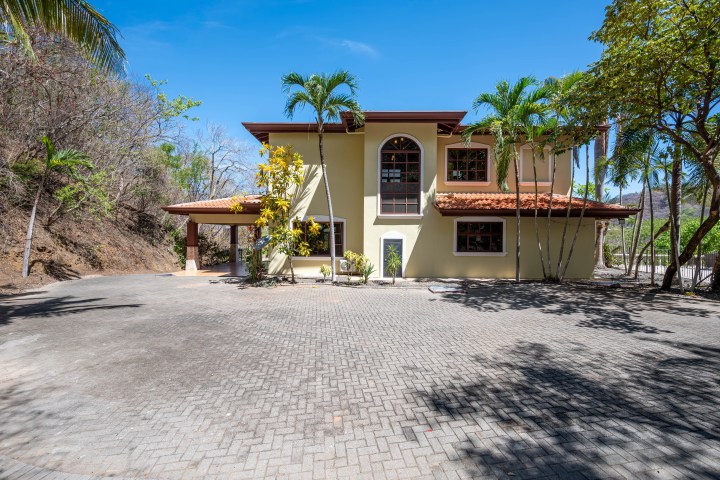 Two-story house with yellow walls, red roof, surrounded by palm trees, clear blue sky.