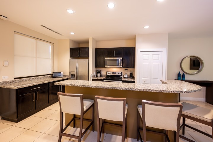 Modern kitchen with granite island, bar stools, stainless steel appliances, and beige tiles.