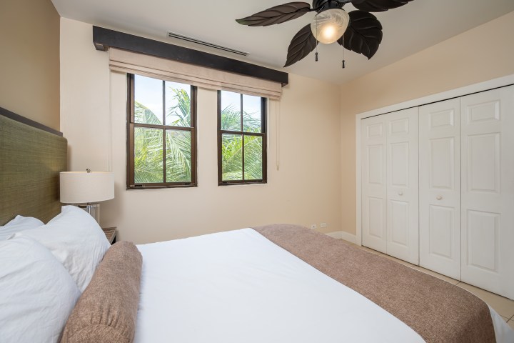 Minimalist bedroom with palm leaf fan, double bed, beige walls, and white closet doors.
