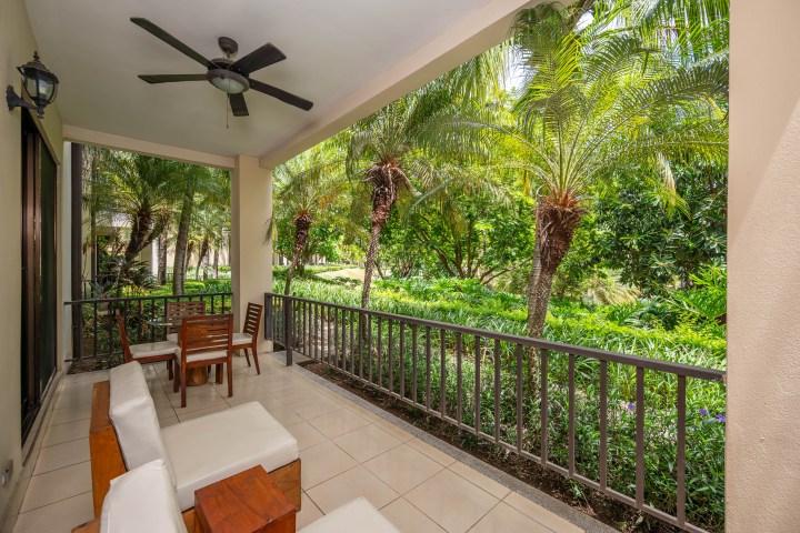 Covered patio with seating, ceiling fan, overlooking lush tropical garden with palm trees.