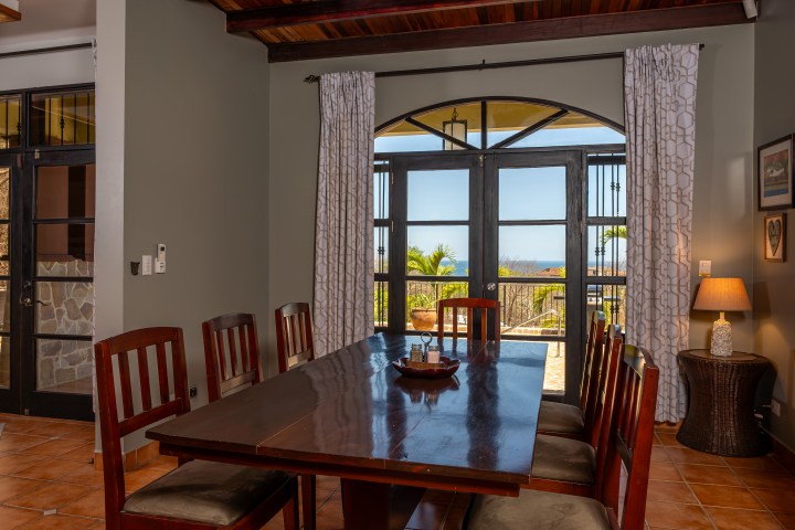 Dining room with wooden table, eight chairs, large window, and view of outdoor greenery.