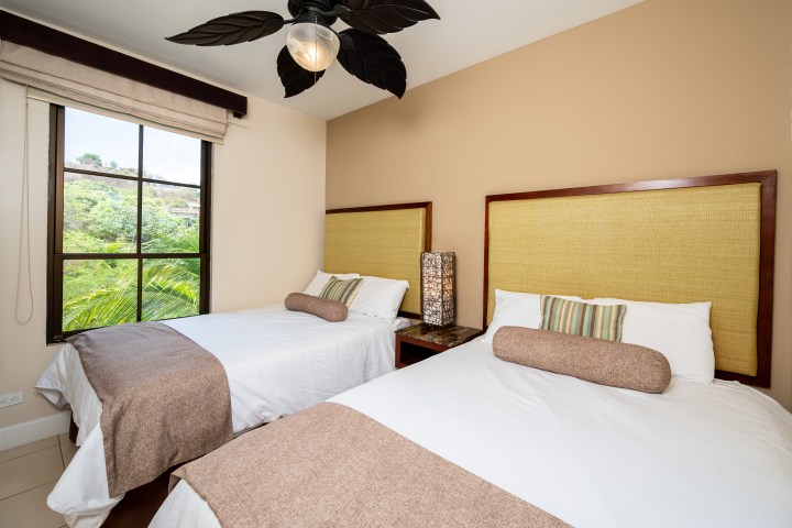 Two beds with beige headboards and a ceiling fan in a bright room.