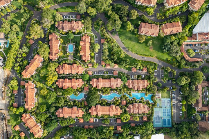 Aerial view of resort with pools, pathways, and red-roofed buildings surrounded by lush greenery.