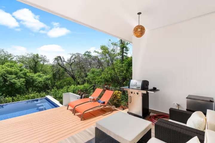 Patio with pool, two red loungers, grill, and outdoor sofa overlooking lush trees.
