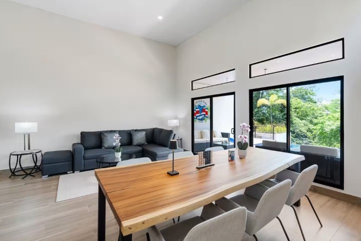 Modern living room with a sectional sofa, wooden table, chairs, and large windows showing greenery.