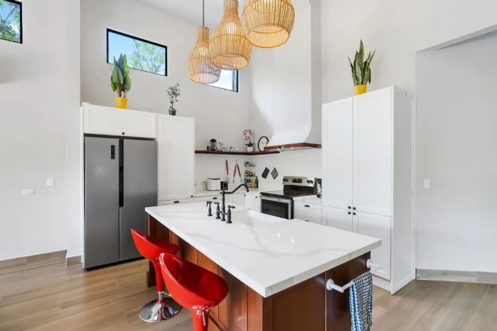 Modern kitchen with island, red stools, pendant lights, and stainless steel appliances.