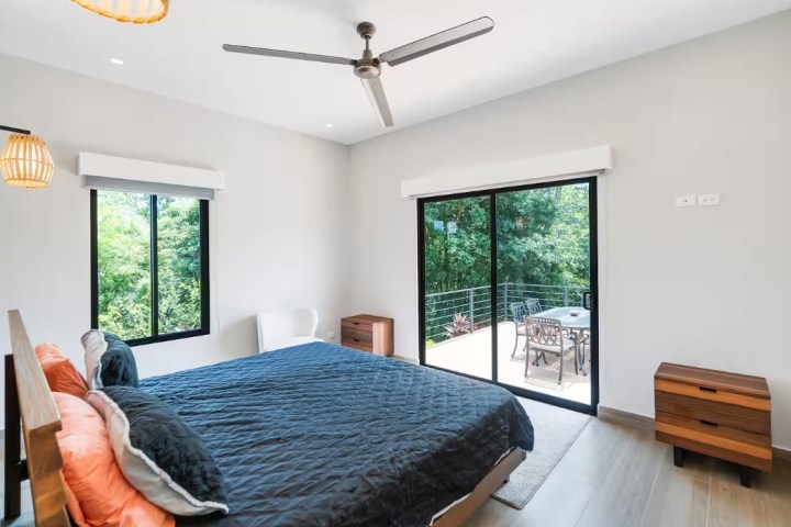 Modern bedroom with bed, ceiling fan, and sliding doors to a balcony overlooking greenery.