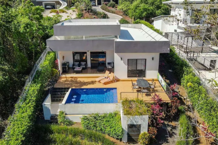 Modern hillside house with pool, surrounded by lush greenery.
