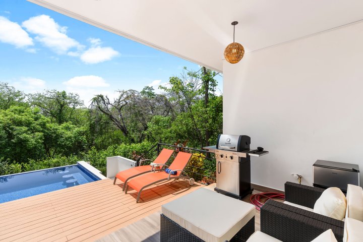 Patio with two orange lounge chairs, BBQ grill, and pool overlooking trees.