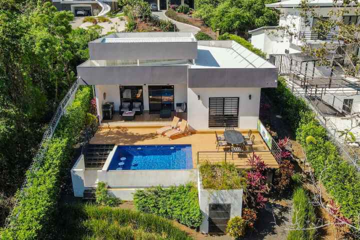 Modern hillside villa with pool, surrounded by lush greenery and a wooden deck.