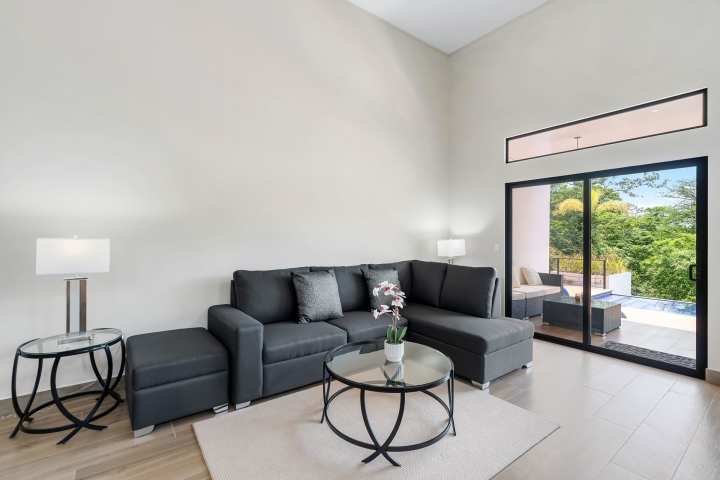 Modern living room with gray sectional sofa, glass tables, and large window view of patio and trees.