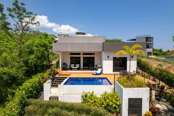 Modern house with pool, surrounded by greenery and distant buildings.