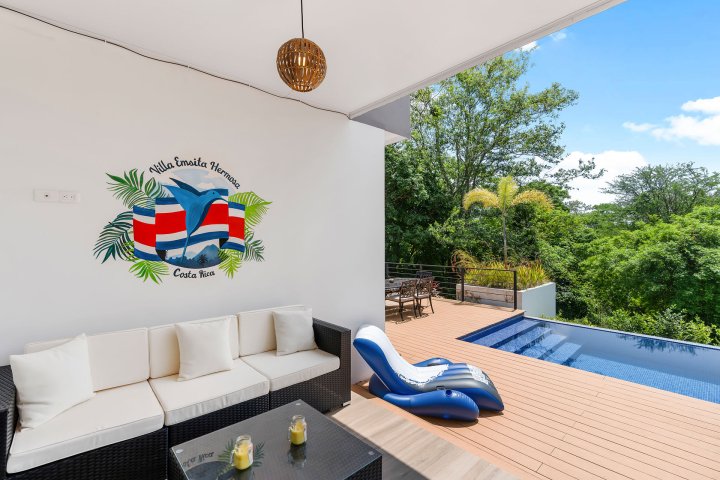 Poolside lounge with white sofa, wall art, and inflated chair by the pool, surrounded by trees.
