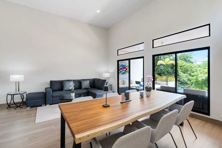 Modern living room with wood dining table, gray sofa, and large windows overlooking greenery.