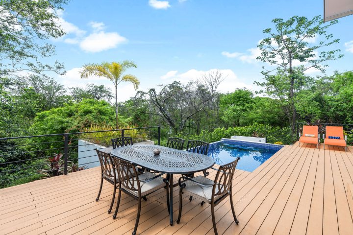Deck with dining set, pool, and trees under a blue sky.