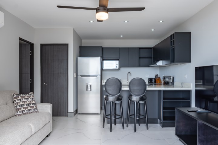 Modern kitchen and living area with sofa, barstools, fridge, and ceiling fan.