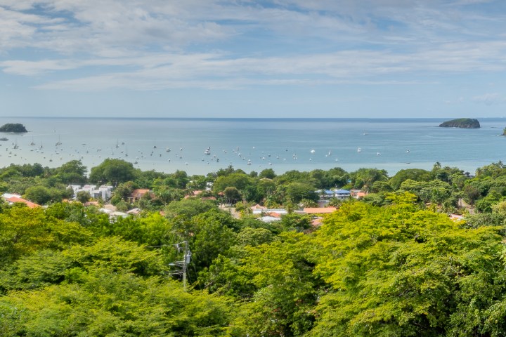 Panoramic view of a coastal town with lush greenery and a distant bay, under a partly cloudy sky.