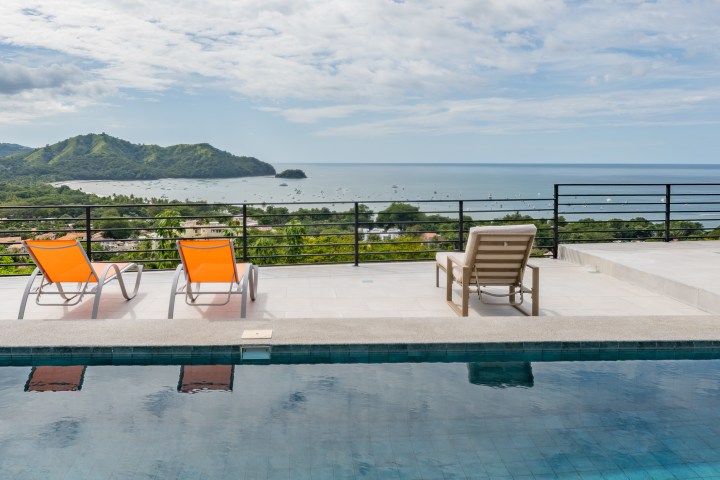 Three lounge chairs by an infinity pool overlook a scenic ocean view.