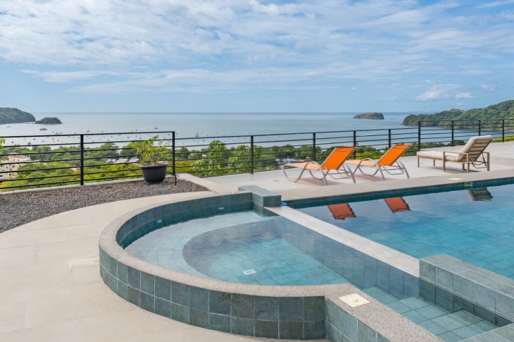 Outdoor pool with ocean view, green hills, and three lounge chairs on sunny day.