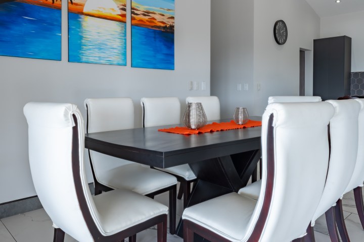 Modern dining room with white chairs and a black table, featuring a triptych painting and orange table runner.