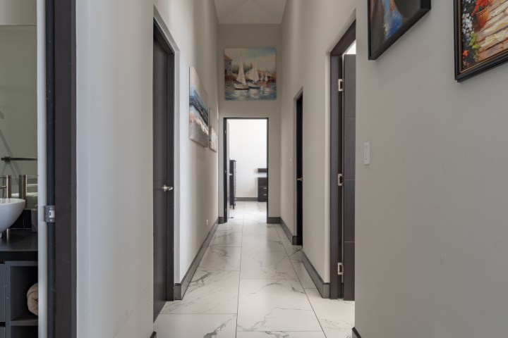 Narrow hallway with white walls, artwork, and doors, leading to a bright room at the end.