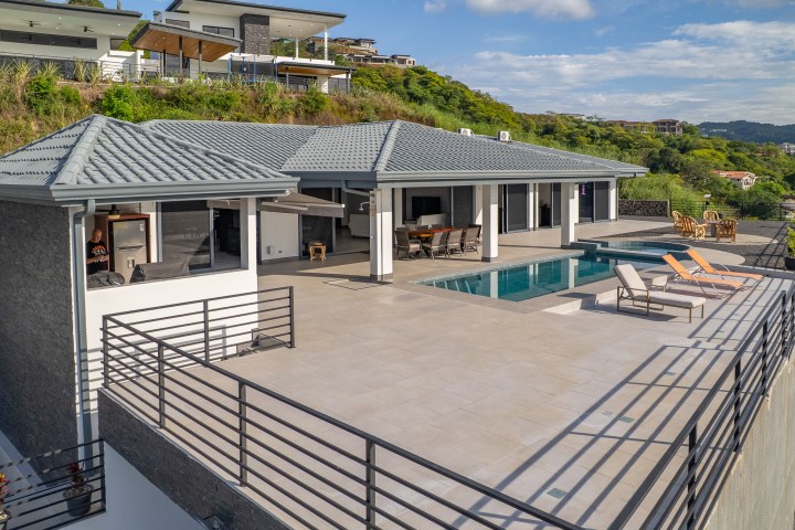 Modern hilltop house with pool and patio overlooking greenery.
