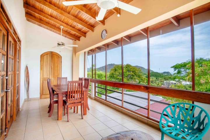 Wooden dining set on balcony with ceiling fan, large windows, and outdoor view.